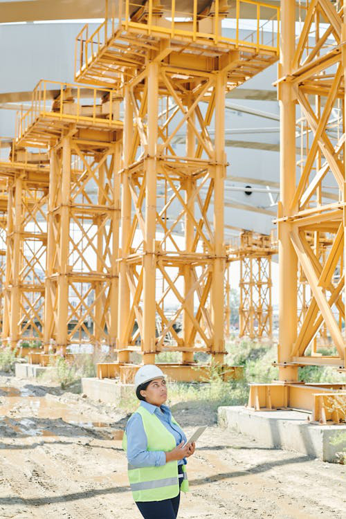 A woman construction worker in a hard hat and vest inspecting yellow steel structures on a building site.