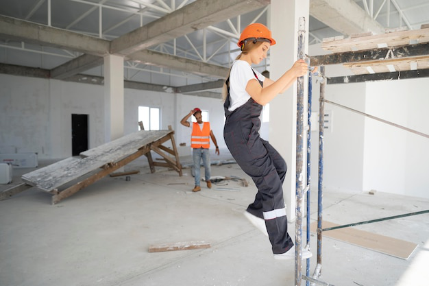 A construction worker wearing a hard hat and safety vest climbs a metal ladder on a busy construction site.