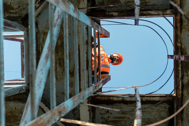 A construction worker wearing a hard hat looks down from a height at a metal ladder on a construction site.