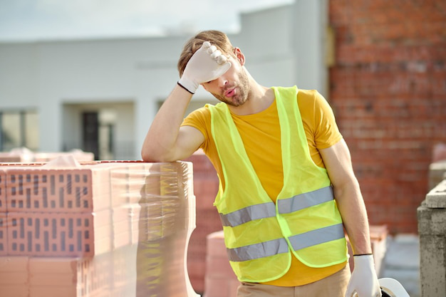 A tired construction worker in a safety vest removes his helmet and rubs his face, standing near a pile of bricks on a sunny construction site.